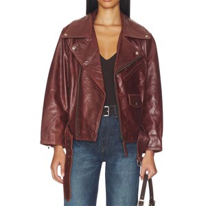 Premium women’s burgundy leather biker jacket featuring an oversized fit, asymmetrical zipper closure, wide lapel collar, vintage finish, adjustable belt detail, and classic moto style paired with denim for a stylish streetwear look.
