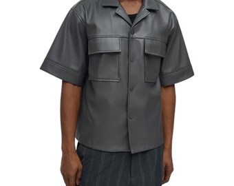 May include: A short-sleeve button-down shirt in a solid dark gray color. The shirt features a classic collar, two chest pockets with flaps, and a smooth finish. It is worn over a black undershirt and paired with dark gray pinstriped trousers.