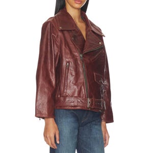 Premium women’s burgundy leather biker jacket featuring an oversized fit, asymmetrical zipper closure, wide lapel collar, vintage finish, adjustable belt detail, and classic moto style paired with denim for a stylish streetwear look.