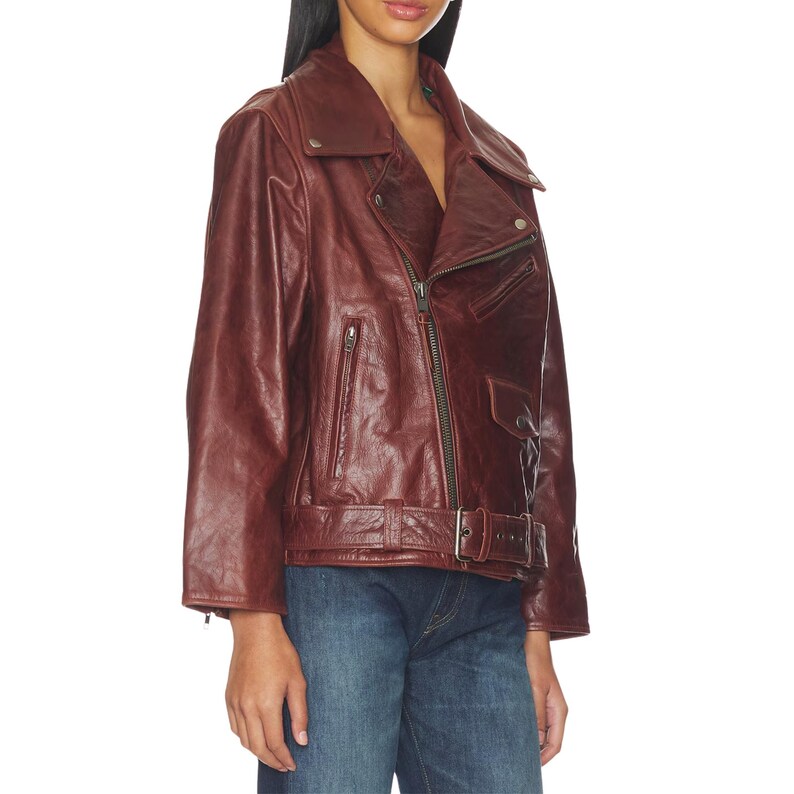 Premium women’s burgundy leather biker jacket featuring an oversized fit, asymmetrical zipper closure, wide lapel collar, vintage finish, adjustable belt detail, and classic moto style paired with denim for a stylish streetwear look.