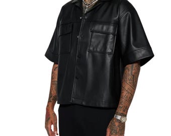 May include: A black short-sleeved shirt made of a smooth, leather-like material. The shirt features a classic collar, a front button-down closure with visible snap buttons, and two symmetrical chest pockets with flaps. The sleeves are wide and straight.