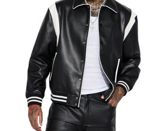 May include: A black faux leather varsity jacket featuring white panels on the shoulders, a pointed collar with white trim, and snap button closures. The cuffs and hem are finished with black and white striped ribbed knit detailing.