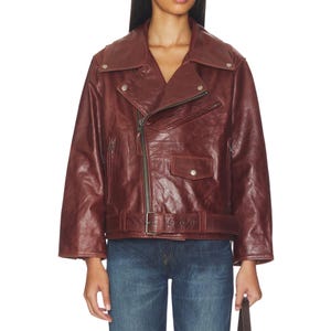 Premium women’s burgundy leather biker jacket featuring an oversized fit, asymmetrical zipper closure, wide lapel collar, vintage finish, adjustable belt detail, and classic moto style paired with denim for a stylish streetwear look.