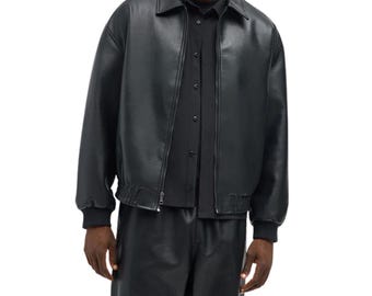 May include: A black leather jacket featuring a classic collar, a full-length front zipper closure, and ribbed cuffs at the wrists and hem. The jacket is worn over a black button-down shirt and paired with matching black leather trousers.