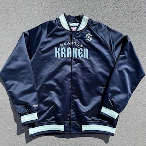 May include: Navy blue satin jacket with the words "SEATTLE KRAKEN" in light blue across the chest. The jacket has a button-up front, a light blue and white striped collar, cuffs, and hem. A white "S" logo is on the right chest.