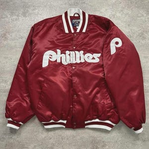 Könnte beinhalten: Bordeauxfarbene Baseballjacke aus Satin mit weißen Akzenten. Die Jacke zeigt den Schriftzug "Phillies" in weißer Schrift auf der Brust und ein weißes "P" auf dem Ärmel. Kragen und Manschetten haben weiße und bordeauxfarbene Streifen.
