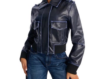 May include: A dark navy blue jacket with a glossy finish, featuring a pointed collar, shoulder epaulets, and two front flap pockets. The jacket has light blue piping along the seams, a black ribbed hem, and black ribbed cuffs, paired with blue jeans.