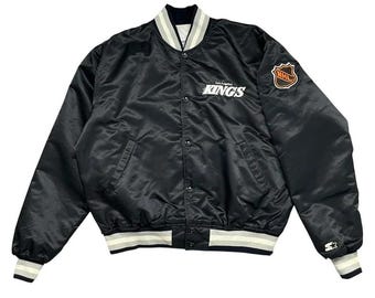 May include: A black satin bomber jacket featuring the Los Angeles Kings logo in white text on the chest, a black and orange NHL shield patch on the left sleeve, a Starter logo on the left cuff, and white and grey striped ribbed trim at the collar and hem.