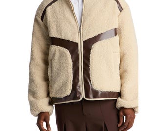 May include: A cream-colored sherpa fleece jacket featuring a full-length front zipper, a high collar, and contrasting dark brown smooth panels across the chest and lower hem. The jacket has long sleeves and a textured, fuzzy exterior finish.