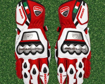 May include: A pair of red, white, and black motorcycle racing gloves featuring the Ducati Corse logo, Italian flag accents, metallic knuckle protectors, and D-TEC engine branding. These protective gloves include reinforced finger padding and wrist closures.