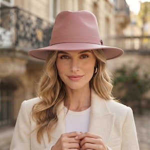 Può includere: Un cappello fedora color rosa polvere con una superficie testurizzata e una fascia tono su tono abbinata attorno alla corona. Il cappello presenta una tesa strutturata e una corona pizzicata, realizzato in materiale solido e opaco.