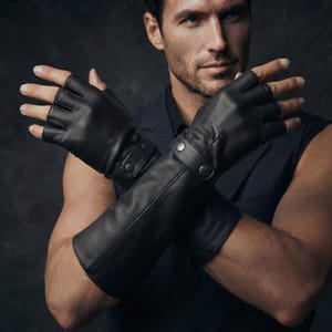 Könnte beinhalten: Ein Paar fingerlose Handschuhe aus schwarzem Leder, die bis zum Unterarm reichen. Die Handschuhe haben eine glatte Textur und werden am Handgelenk mit einem Riemen befestigt, der zwei silberfarbene Metalldruckknöpfe aufweist. Die Finger sind frei.