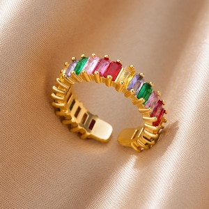 May include: A gold-toned open ring featuring a row of rectangular baguette-cut stones in various colors including red, green, yellow, purple, and pink. The band has a polished metallic finish and an adjustable gap at the bottom for sizing.