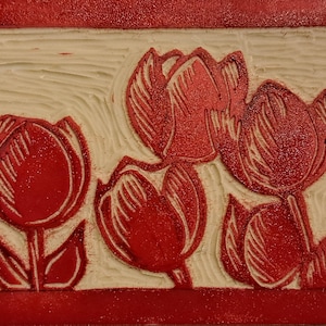 Op de afbeelding: Een rechthoekig kunstwerk met een gesneden ontwerp van rode tulpen op een crèmekleurige achtergrond. De tulpen hebben gedetailleerde bloemblaadjes en stelen, geplaatst binnen een rode glitterrand. Het stuk heeft een getextureerde, handgemaakte uitstraling.