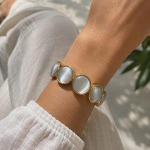 May include: A gold-toned bracelet featuring a series of circular, smooth, translucent white cabochon stones. The stones are set in polished gold metal frames and linked together to form a continuous band that encircles the wrist.