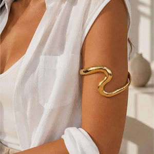 May include: A polished gold-tone arm cuff featuring a continuous, fluid, wavy design that wraps around the upper arm. The metal surface is smooth and reflective, creating a bold, sculptural shape that sits securely on the bicep area.