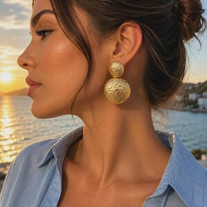 May include: A pair of gold-toned earrings featuring two stacked spheres with a textured, hammered finish. The smaller sphere sits at the top, while the larger sphere hangs below, creating a bold, geometric design with a metallic luster.