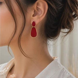 May include: A pair of drop earrings featuring two stacked, teardrop-shaped red enamel components encased in thin gold-toned metal frames. The smaller teardrop sits above the larger one, creating a vertical, layered design with a smooth finish.