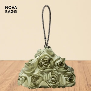 Op de afbeelding: Een saliegroene clutch met een rozenachtig ontwerp. De tas heeft een kettinghandvat en de tekst "NOVA BAGG" in de linkerbovenhoek. De tas is gemaakt van een zacht, getextureerd materiaal.