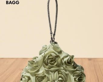 May include: A sage green clutch bag with a rose-like design. The bag features a chain handle and the text "NOVA BAGG" in the upper left corner. The bag is made of a soft, textured material.
