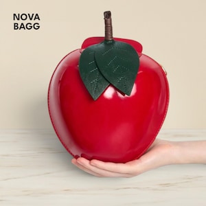 Könnte beinhalten: Eine glänzende, rot geformte Handtasche in Apfelform mit grünen Blättern und einem braunen Stiel. Die Tasche wird in einer Hand gehalten. Der Text "NOVA BAGG" befindet sich in der oberen linken Ecke.