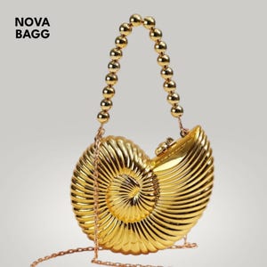 Könnte beinhalten: Eine goldfarbene Handtasche in Form eines Nautilus-Muschelschalen. Sie hat einen Perlenhenkel und einen feinen Kettenriemen. Der Text "NOVA BAGG" befindet sich in der oberen linken Ecke.