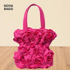 Puede incluir: Un bolso de mano rosa brillante con asa, cubierto de rosas de tela. El bolso tiene forma rectangular y el texto "NOVA BAGG" está impreso en la esquina superior izquierda.
