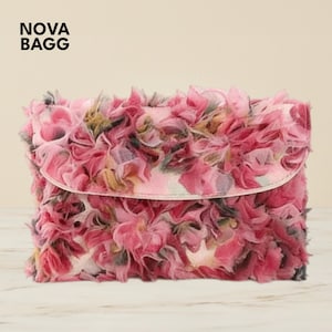 Op de afbeelding: Een roze clutch met een bloemenontwerp. De tas is bedekt met lagen roze stof, waardoor een getextureerd, gerimpeld effect ontstaat. De flap is een lichtere tint roze. De tekst "NOVA BAGG" staat linksboven.