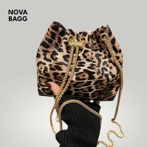 Puede incluir: Un bolso tipo cubo con estampado de leopardo y correa de cadena dorada. El bolso es marrón y negro con cierre de cordón y herrajes dorados. El texto "NOVA BAGG" está en la esquina superior izquierda.
