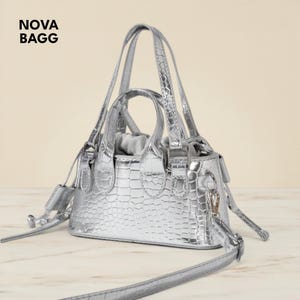 Può includere: Una borsa argentata con una texture a effetto pelle di coccodrillo. La borsa ha due manici corti e una lunga tracolla staccabile. Il testo "NOVA BAGG" è nell'angolo in alto a sinistra.