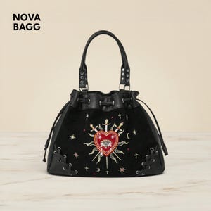 Può includere: Borsa nera con coulisse e design ricamato. Il design presenta un cuore rosso con un occhio, circondato da spade, stelle e lune crescenti. La borsa ha un manico superiore e il testo "NOVA BAGG".