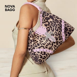 Peut inclure: Un sac à main à imprimé léopard avec des détails roses et une croix argentée. Le sac a une poignée et des bordures rose clair, avec des clous argentés. Le texte "NOVA BAGG" est visible.