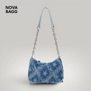Puede incluir: Un bolso de hombro de mezclilla con asa azul claro y correas de cadena plateadas. El bolso presenta un diseño de patchwork con patrones de estrellas. El texto "NOVA BAGG" es visible en la esquina superior izquierda.