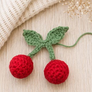 Peut inclure: Une cerise au crochet faite à la main, composée de deux sphères rouges et de feuilles vertes. La cerise est posée sur une surface en bois clair, avec un article tricoté crème en arrière-plan. Un objet décoratif.