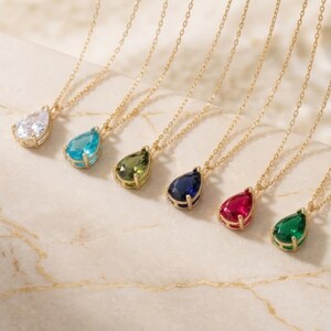 May include: Six gold necklaces, each featuring a single pear-shaped pendant. The pendants include a clear stone, a light blue stone, an olive green stone, a dark blue stone, a red stone, and an emerald green stone, all set in gold prongs.