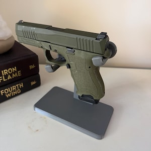 Könnte beinhalten: Ein olivgrünes halbautomatisches Pistolenmodell auf einem grauen Kunststoff-Präsentationsständer. Die Schusswaffe verfügt über einen strukturierten Griff, einen Schlitten mit gezackten hinteren Kanten, ein Korn und einen schwarzen Magazinboden.