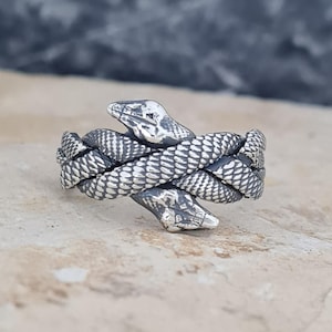 Può includere: Un anello in argento raffigurante due serpenti intrecciati con squame dettagliate. Le teste dei serpenti sono rivolte in direzioni opposte, formando la fascia dell'anello. Il metallo presenta una finitura lucida e una texture scura.