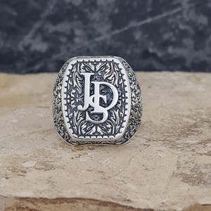 May include: A silver-toned signet ring featuring an intricate, dark-oxidized floral or vine-like relief pattern across the band and the rectangular face. The center of the ring displays the prominent, raised, polished silver letters J and D.