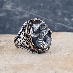Peut inclure: Une bague en argent avec une incrustation de pierre noire ovale centrée sur un cobra en argent tridimensionnel détaillé. L'anneau est orné de motifs en filigrane d'argent complexes et accentué par de petites perles métalliques dorées sur le bord.