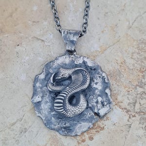 Op de afbeelding: Een zilverkleurige hanger aan een ketting met een reliëf van een opgerolde cobra op een ronde, getextureerde donkere metalen schijf. De hanger hangt aan een bijpassende zilverkleurige schakelketting met een ruwe, gehamerde afwerking op het oog.