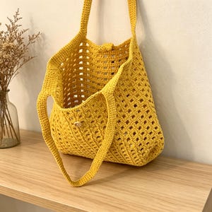 Könnte beinhalten: Eine sonnengelbe Häkeltasche mit Knopfverschluss. Die Tasche hat zwei Henkel und ein rechteckiges, offenes Webmuster. Die Tasche liegt auf einer Holzoberfläche.