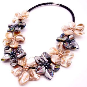 May include: A statement necklace featuring a black braided cord and a series of floral-shaped clusters made from iridescent, multi-colored shell petals. The centers of the flowers are accented with small, dark, round beads and metallic spheres.