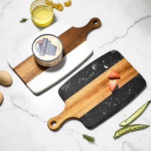 May include: Two rectangular serving boards with handles, one featuring white marble and wood, the other black marble and wood. The white board holds a round container of Ile de France Brie au Bleu cheese. Two strawberries rest on the black board.