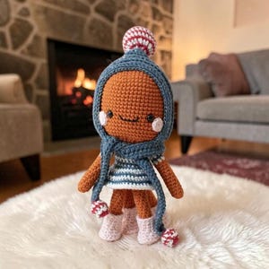 Könnte beinhalten: Eine gehäkelte Puppe mit rundem orangefarbenem Kopf, schwarzen Augen und rosa Wangen. Sie trägt eine blaue Kapuzenmütze mit rot-weißem Bommel, einen passenden blauen Schal mit Bommeln, ein blau-weiß gestreiftes Oberteil und hellrosa gehäkelte Stiefel.