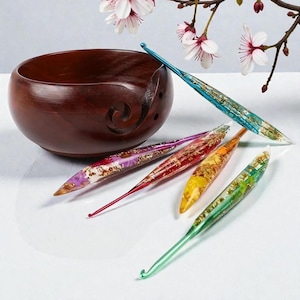 May include: A dark brown wooden yarn bowl with a spiral cutout alongside five resin crochet hooks in blue, purple, red, yellow, and green, each containing embedded gold leaf flakes and dried flower petals arranged on a white surface.