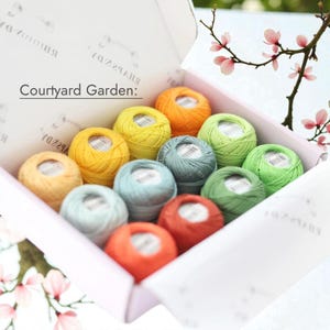 Peut inclure: Une boîte en carton blanc contenant douze petites pelotes de fil de différentes couleurs, notamment jaune, orange, vert et bleu. Le couvercle intérieur de la boîte porte le texte Courtyard Garden et la marque Rhapsody imprimés en noir.