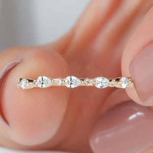 May include: A gold band ring featuring a series of alternating pear-shaped and round clear stones set along the top. The metal has a warm yellow tone, and the stones are arranged in a linear pattern across the front of the thin, delicate band.