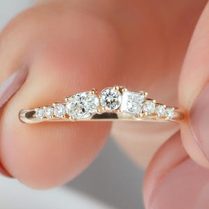 May include: A gold ring featuring a curved band set with a variety of clear, faceted stones in different shapes, including round, oval, and square cuts, arranged in a cluster across the top of the band, creating a delicate and symmetrical design.
