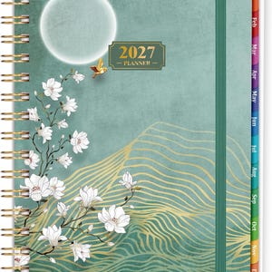 May include: A 2027 planner with a sage green cover featuring a white moon, a small gold bird, and white magnolia flowers. The design includes gold wavy lines, a gold rectangular label reading 2027 PLANNER, a gold spiral binding, and monthly tabs.