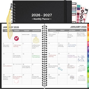 May include: Black spiral-bound monthly planner for 2026-2027. The open pages show a January 2026 calendar with handwritten notes and stickers. Includes a ribbon bookmark and a set of colorful sticker tabs.